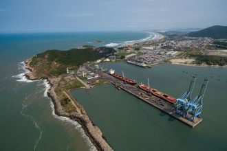 Vista aérea do porto de Imbituba, com navios, cais e guindastes junto à costa brasileira.