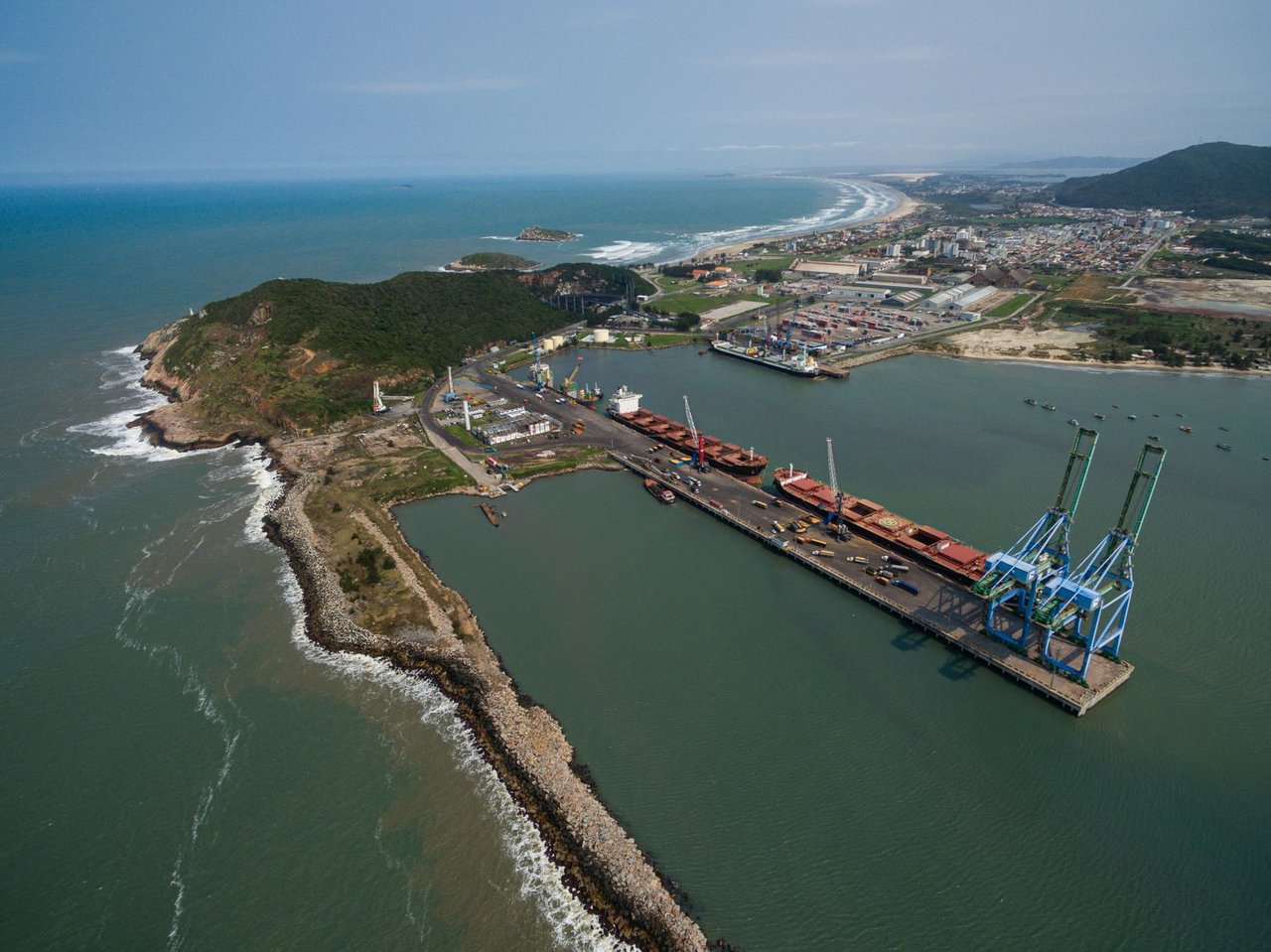 Vista aérea do porto de Imbituba, com navios, cais e guindastes junto à costa brasileira.